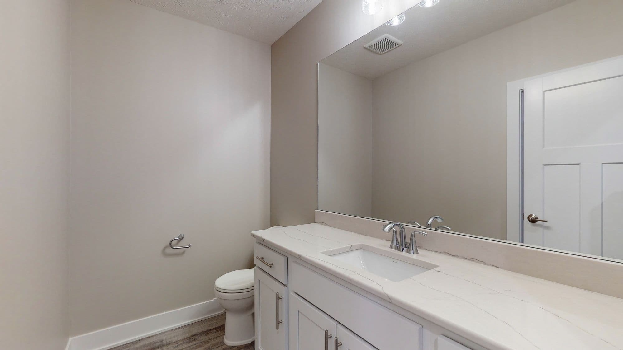 Bathroom featuring a vanity with a white countertop and cabinetry, chrome fixtures, a toilet, and a large mirror reflecting the room. The walls are painted in a neutral tone, complemented by the white trim and the light wood-look flooring.