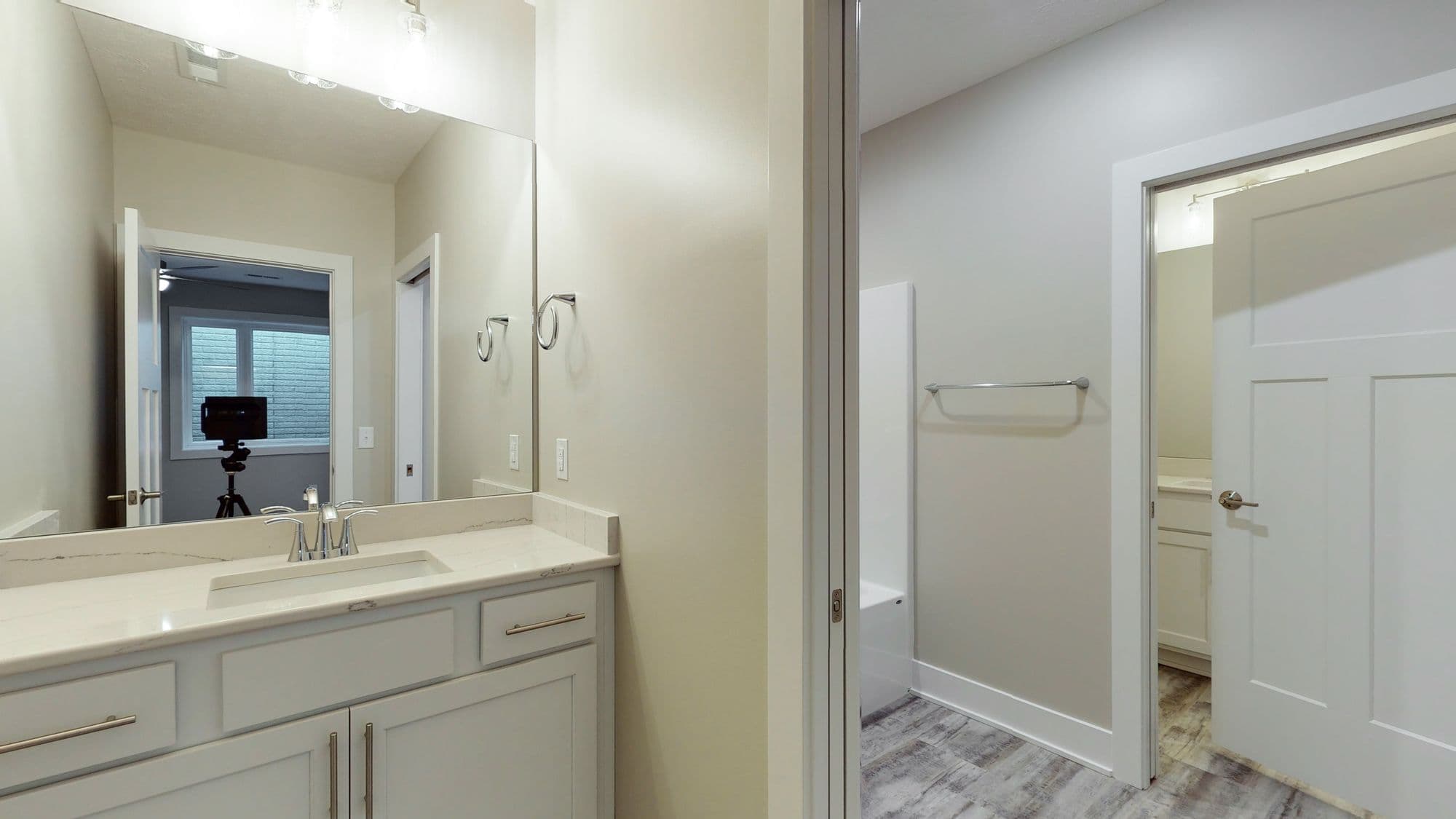 Bathroom featuring a white vanity with marble countertop, chrome fixtures, and a large mirror. A doorway leads to a separate room with a shower and gray wood-look flooring.