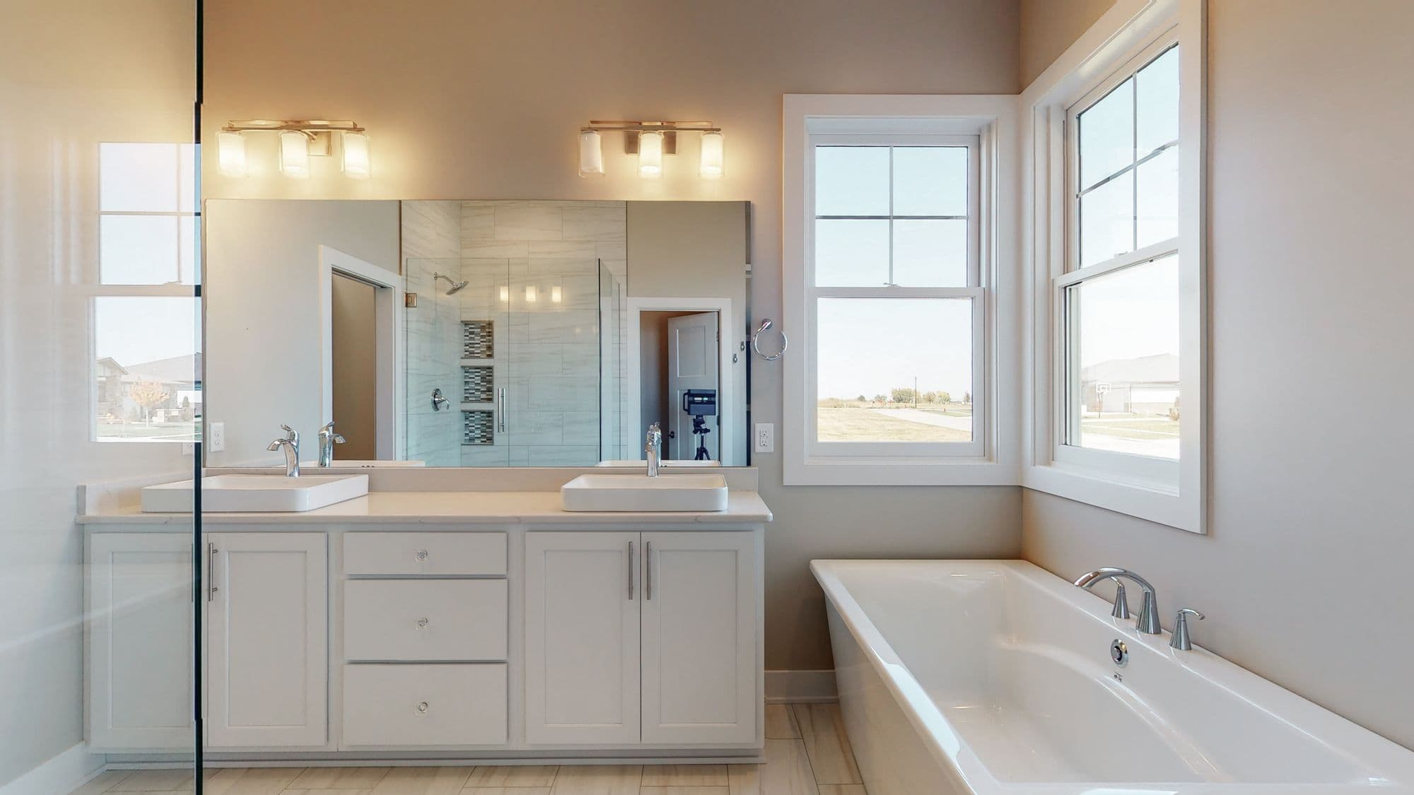 Modern bathroom with double vanity, white cabinets, and quartz countertops. Large mirror and window with white trim add light to the neutral-toned space.