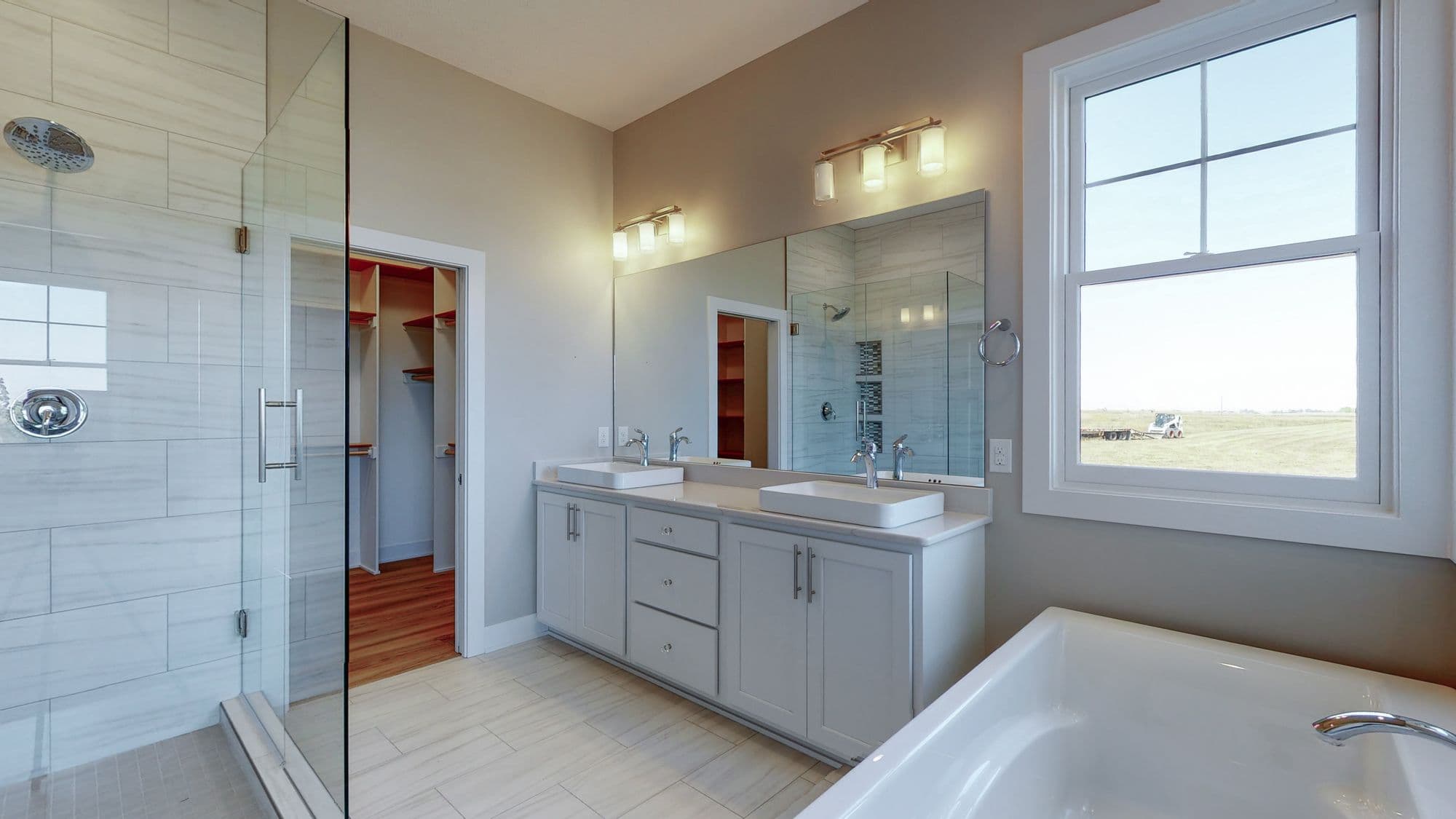 Modern bathroom features a double vanity with white countertops and gray cabinetry. A glass-enclosed shower is visible to the left, while a large window and soaking tub are located to the right, providing natural light.