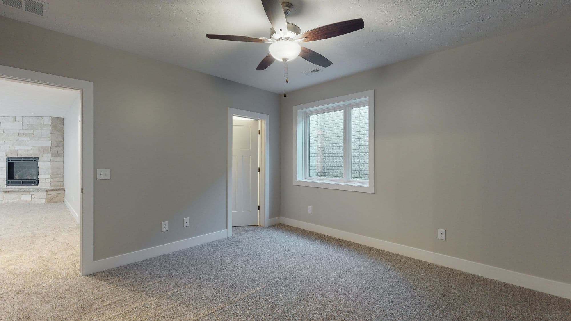 Bedroom features neutral walls, gray carpet, and a ceiling fan. A window well provides natural light, while a doorway leads to another room with a stone fireplace.