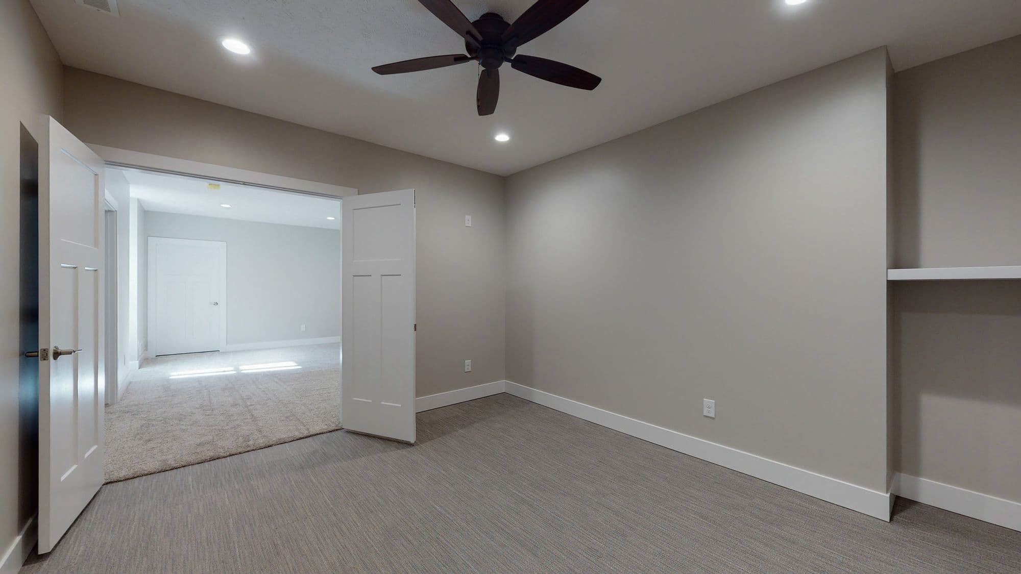 This is a bedroom with gray carpet and neutral-toned walls, featuring a ceiling fan and white trim. An open white door leads to another carpeted room.