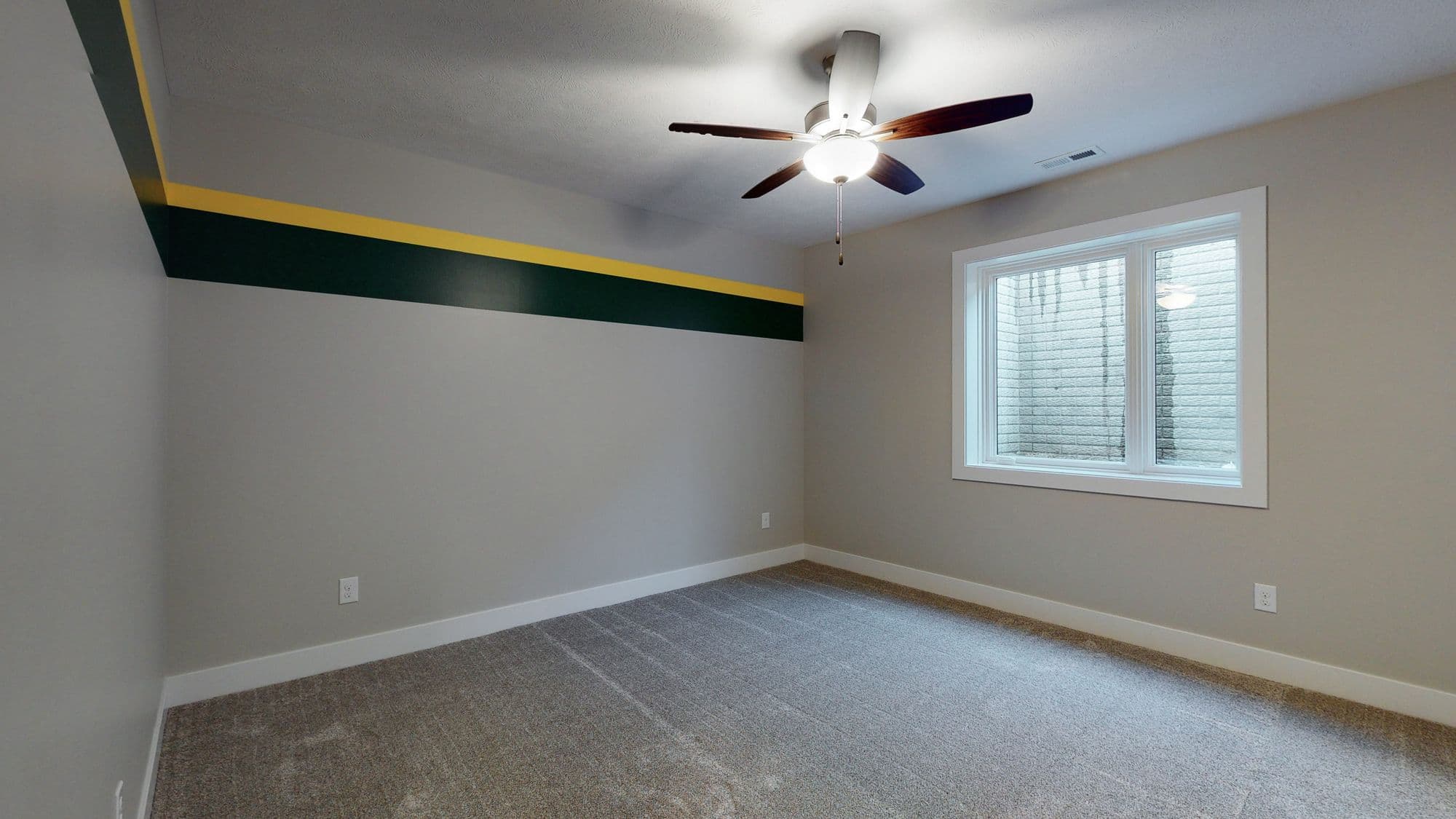 Bedroom interior features a neutral color palette with cool gray walls complemented by a gray carpet. A decorative green and yellow stripe accents the upper wall, and a ceiling fan with dark wooden blades provides air circulation.