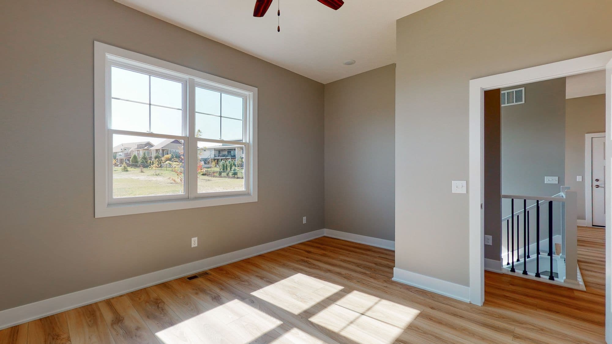 A brightly lit bedroom showcases a large window offering a view of the outside neighborhood. The room features wood flooring and neutral-toned walls, leading to an open doorway with a view of the upper level.