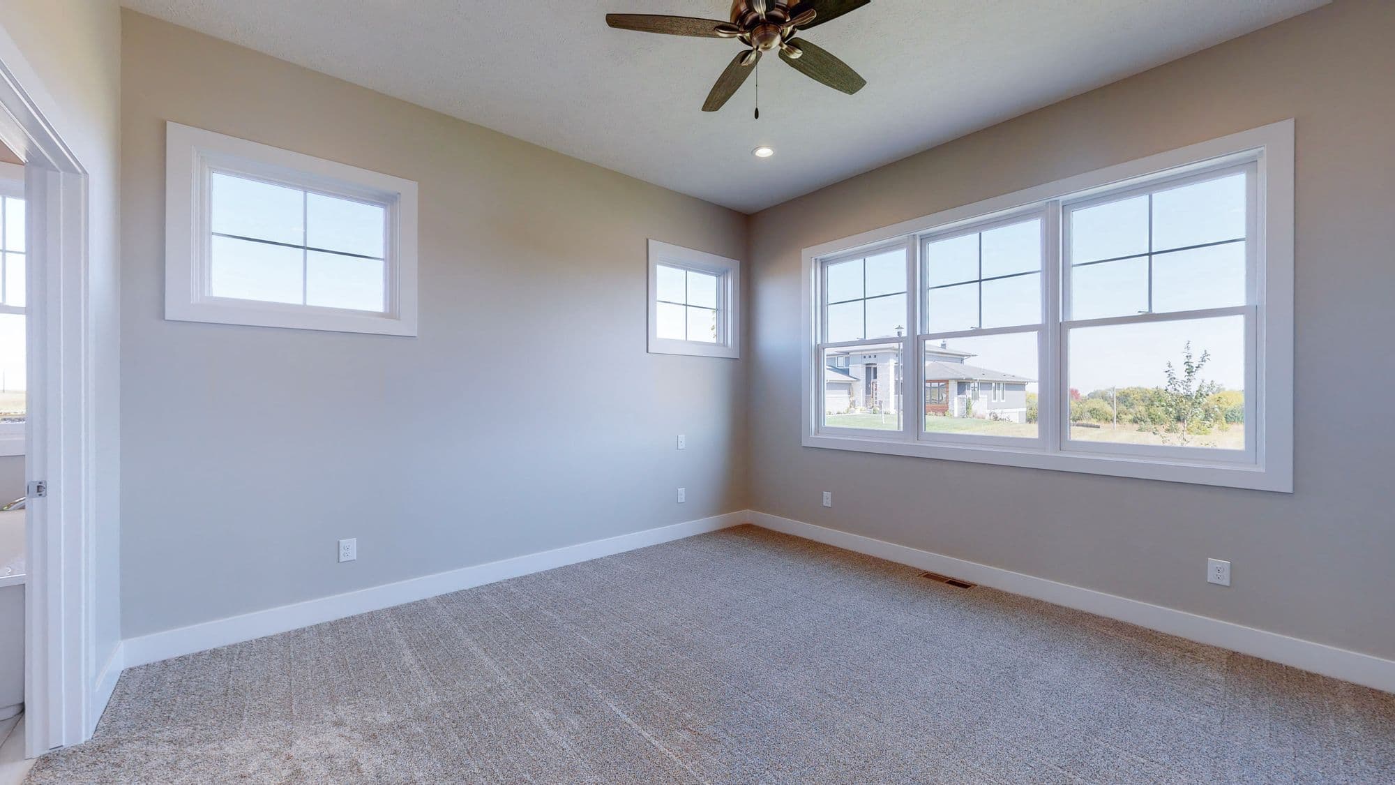 The room is an empty bedroom with a neutral color palette, featuring three windows that provide natural light. There is carpeted flooring and a ceiling fan in the center of the room.