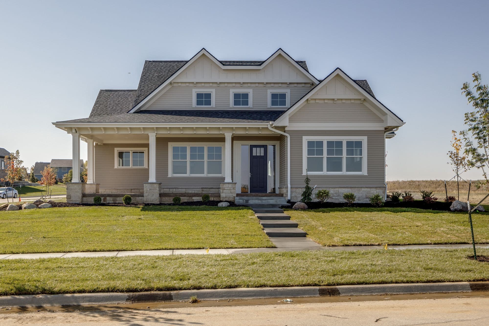 Exterior view of a single-story home with light brown siding, a dark gray roof, and white trim. The home features a covered front porch with white columns and a well-manicured lawn.