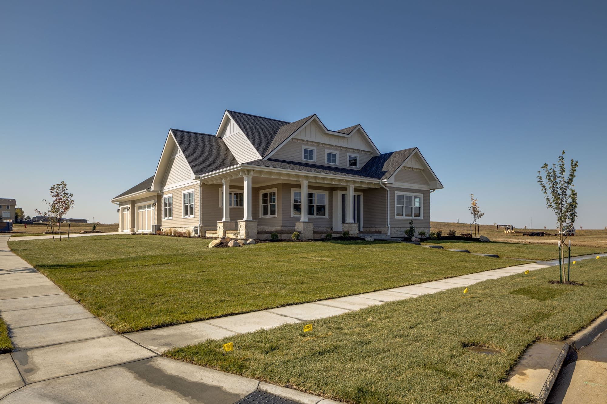 New construction home with light beige siding, gabled roof, and a covered front porch supported by stone columns. Manicured lawn and concrete sidewalk lead to the house.