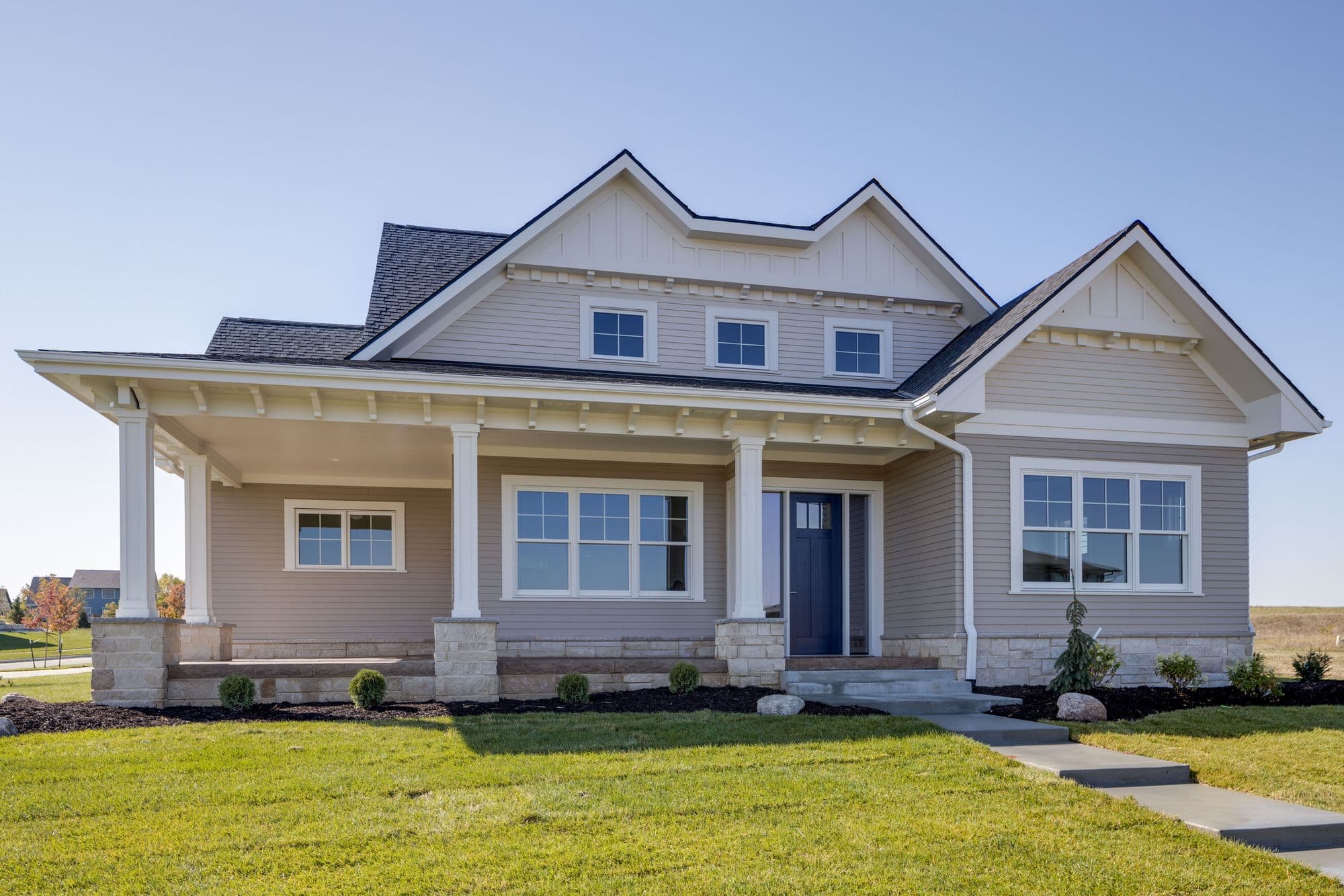 Exterior view of a light beige house with a covered front porch supported by white columns with stone bases. The house features a dark roof, white trim, and well-manicured lawn and landscaping.