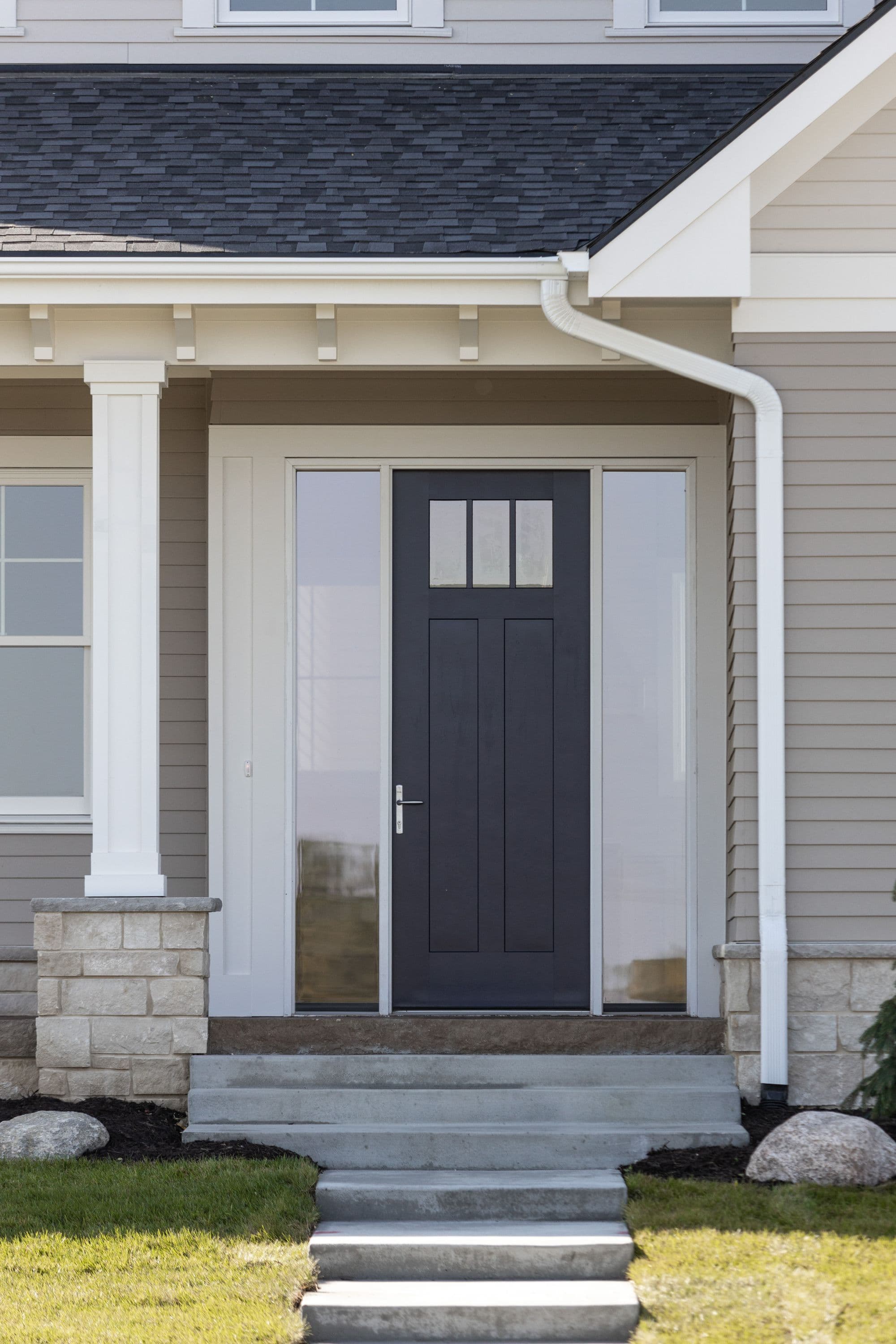 Exterior view showcasing a home's entrance featuring a dark gray front door with sidelights, complemented by a concrete porch with steps. The house is clad in light gray siding, with white trim and a dark gray roof.
