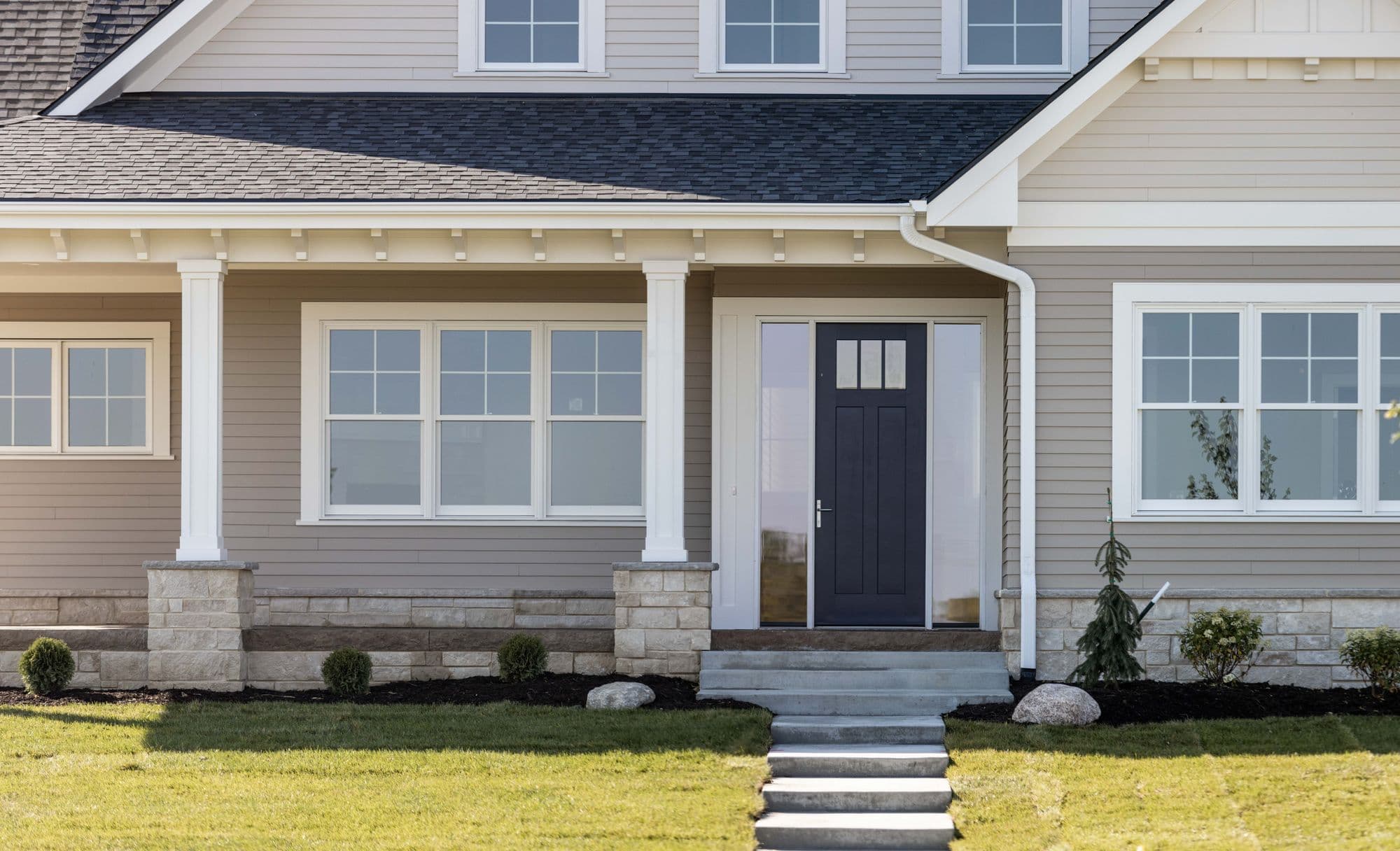 Front exterior of a two-story home featuring horizontal lap siding, a dark blue front door with sidelights, a covered porch supported by white columns with stone bases, and well-maintained landscaping.