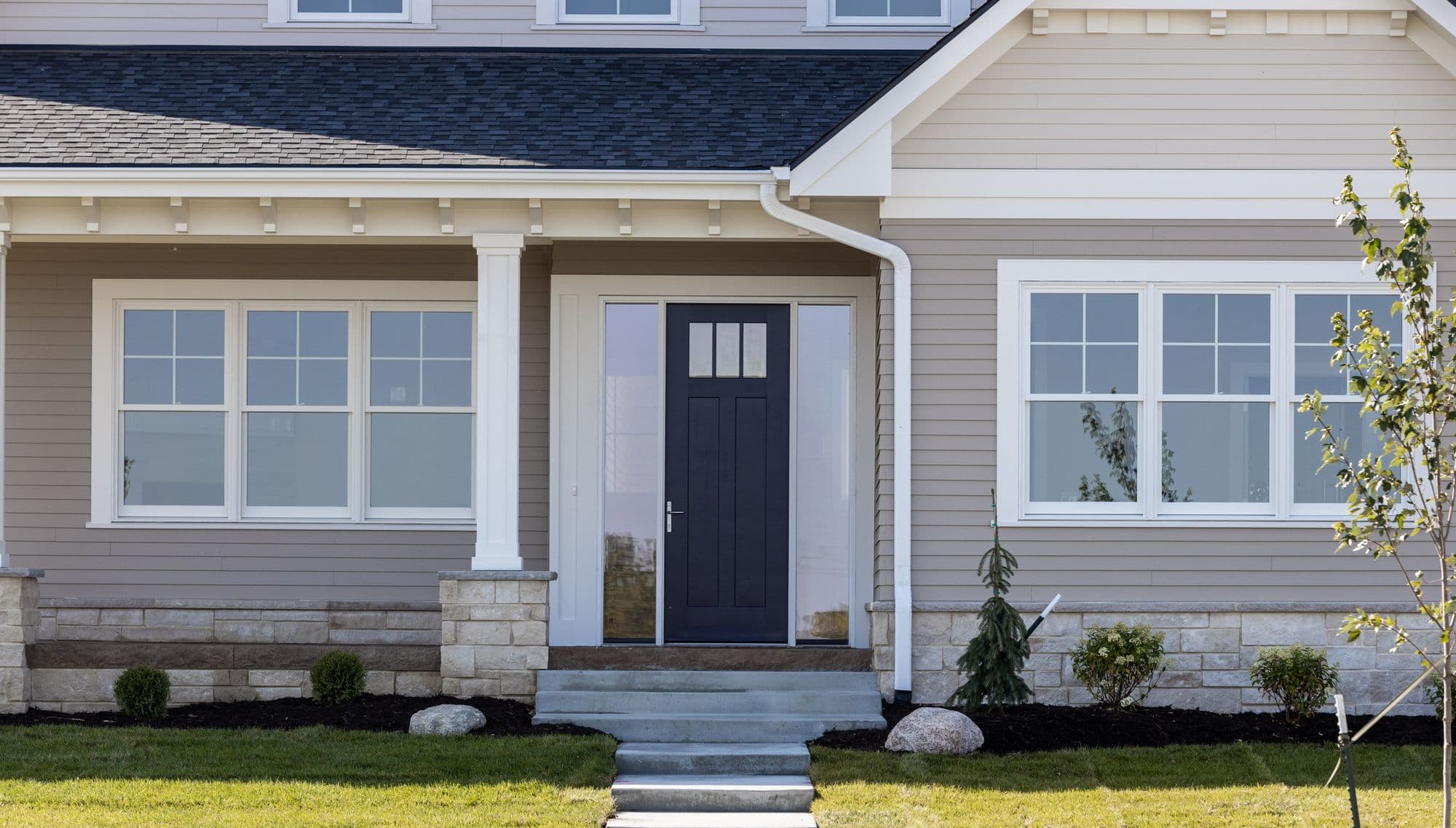 Exterior shot of a house with a dark blue front door framed by sidelights and flanked by white-framed windows. The facade features light gray lap siding with stone accents at the base, complemented by a dark gray roof and a well-manicured lawn.