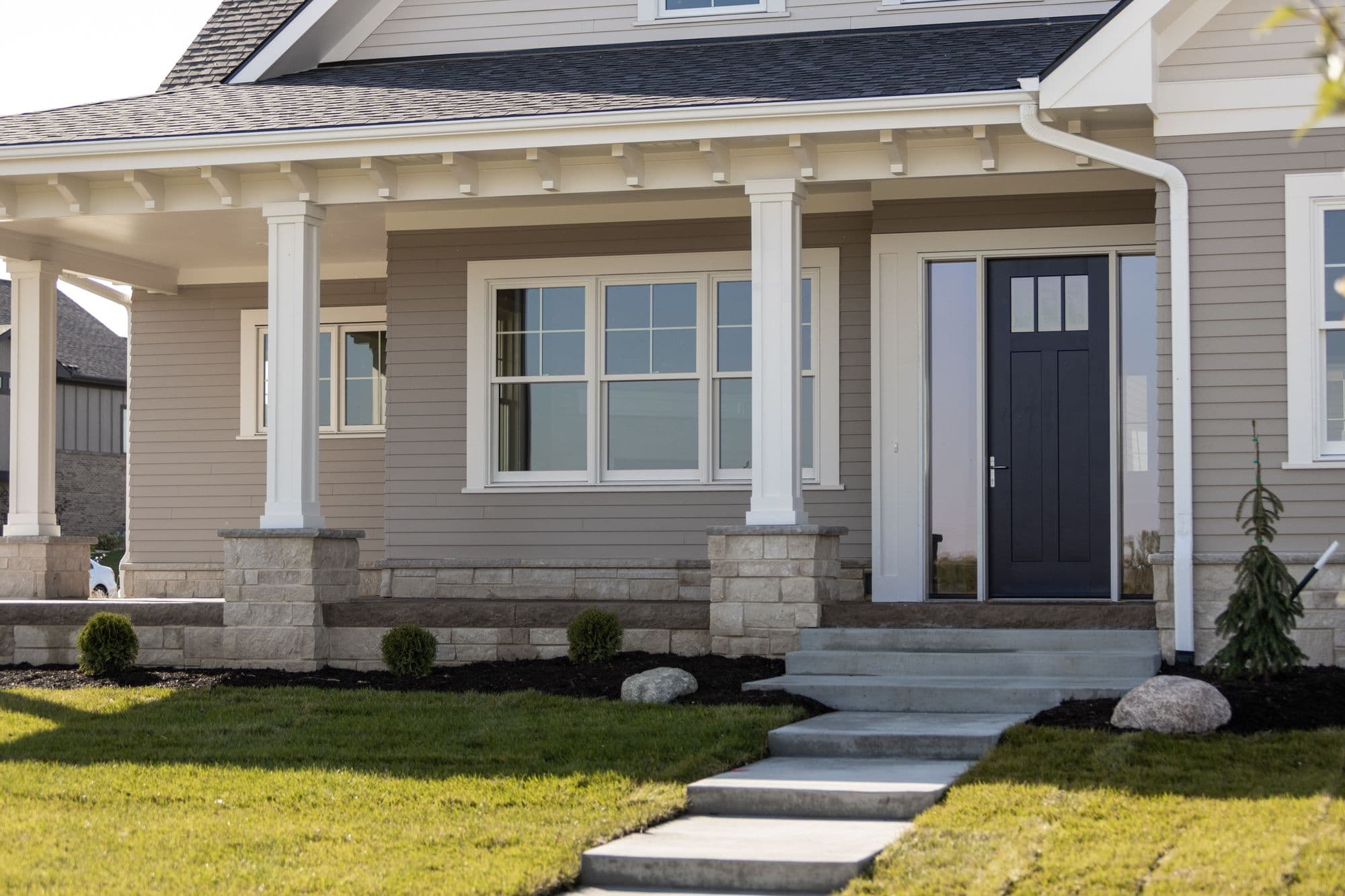 Tan home exterior features a covered porch with white columns and stone accents, a dark blue front door with glass panels, and a well-manicured lawn with stone accents. Steps lead to the front door.