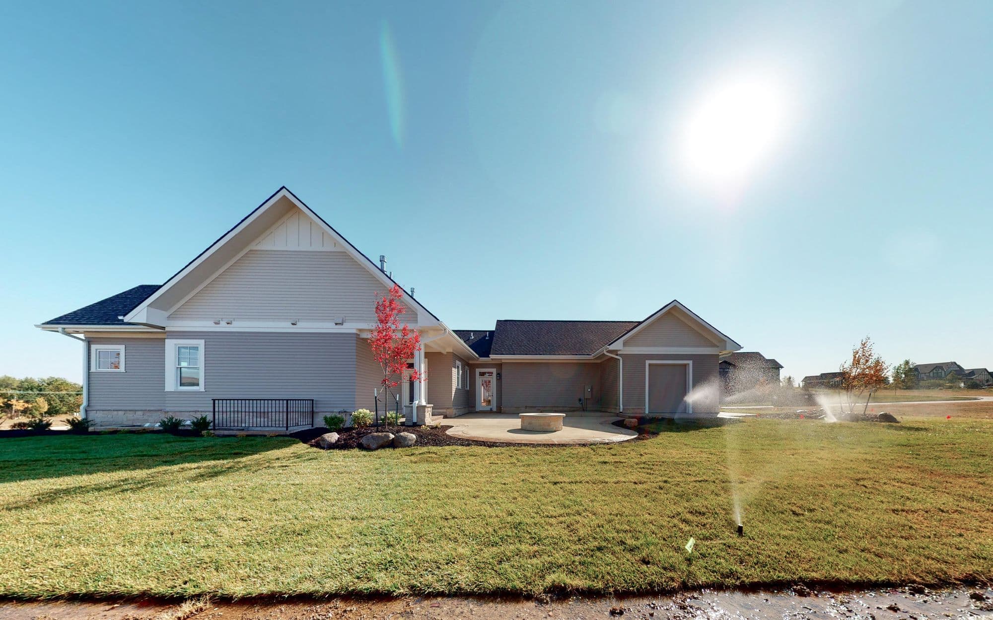 Exterior of a modern single-family home with light gray siding, a dark roof, and manicured green lawn. Features include a concrete patio with a fire pit and a small red-leaved tree.