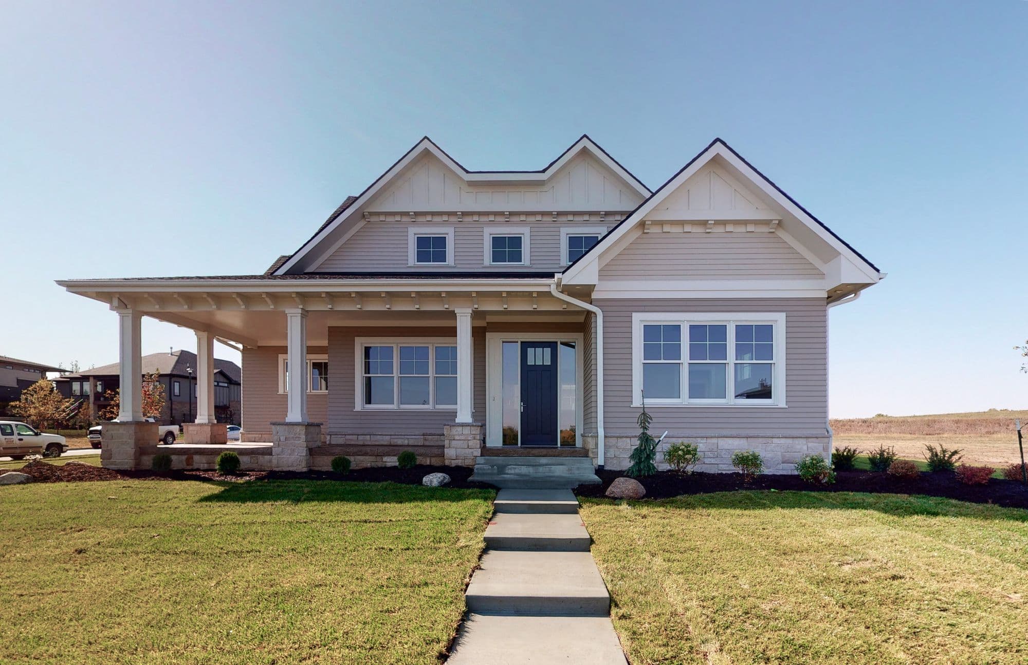 Exterior view of a two-story home featuring a covered front porch with stone columns, light gray lap siding, and multiple windows. A concrete walkway leads to the dark blue front door.