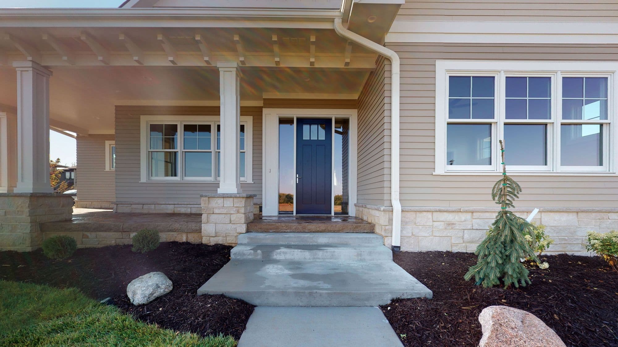 The image shows the exterior of a house featuring a covered front porch with stone-based columns and a dark blue front door. A large window with white trim is visible to the side, and landscaping includes a small tree and various ground cover.