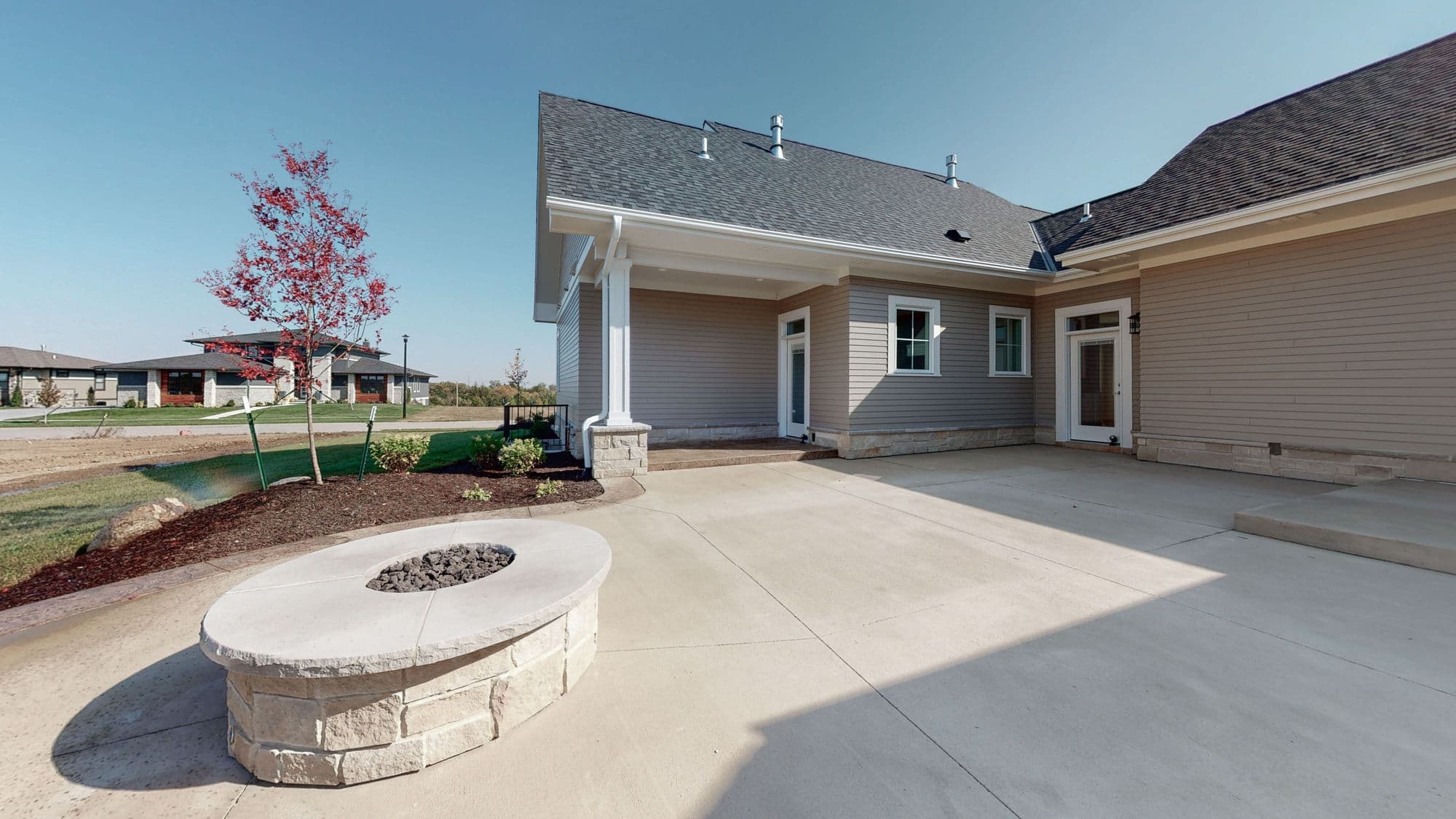 Exterior view of a modern home with a concrete patio featuring a stone fire pit. The house has light gray siding, white trim, and a dark gray roof.