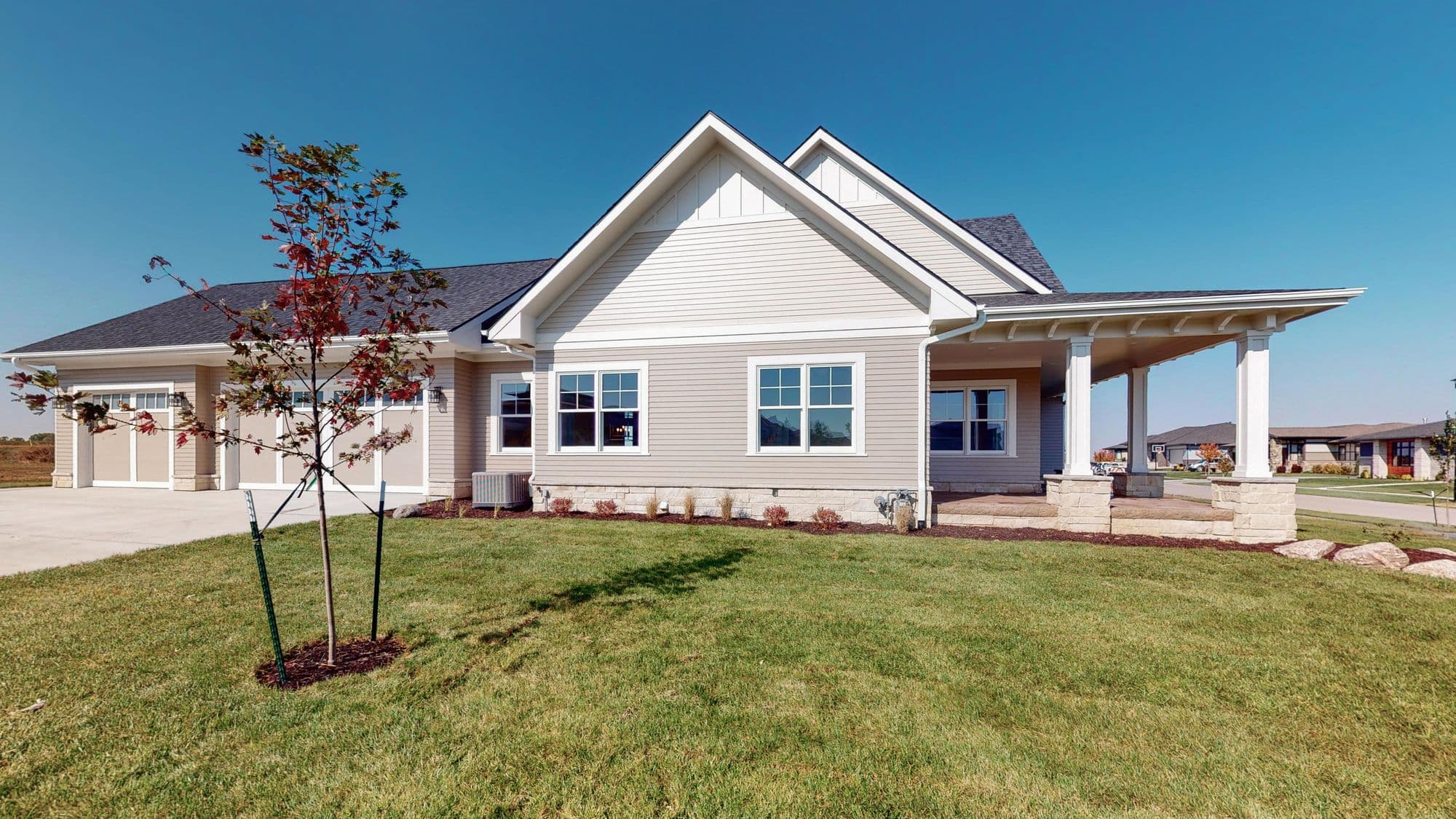 Residential exterior featuring a home with a gable roof, lap siding, and stone veneer details. A young tree in the front yard adds a touch of greenery, and a covered porch extends from the side of the house.