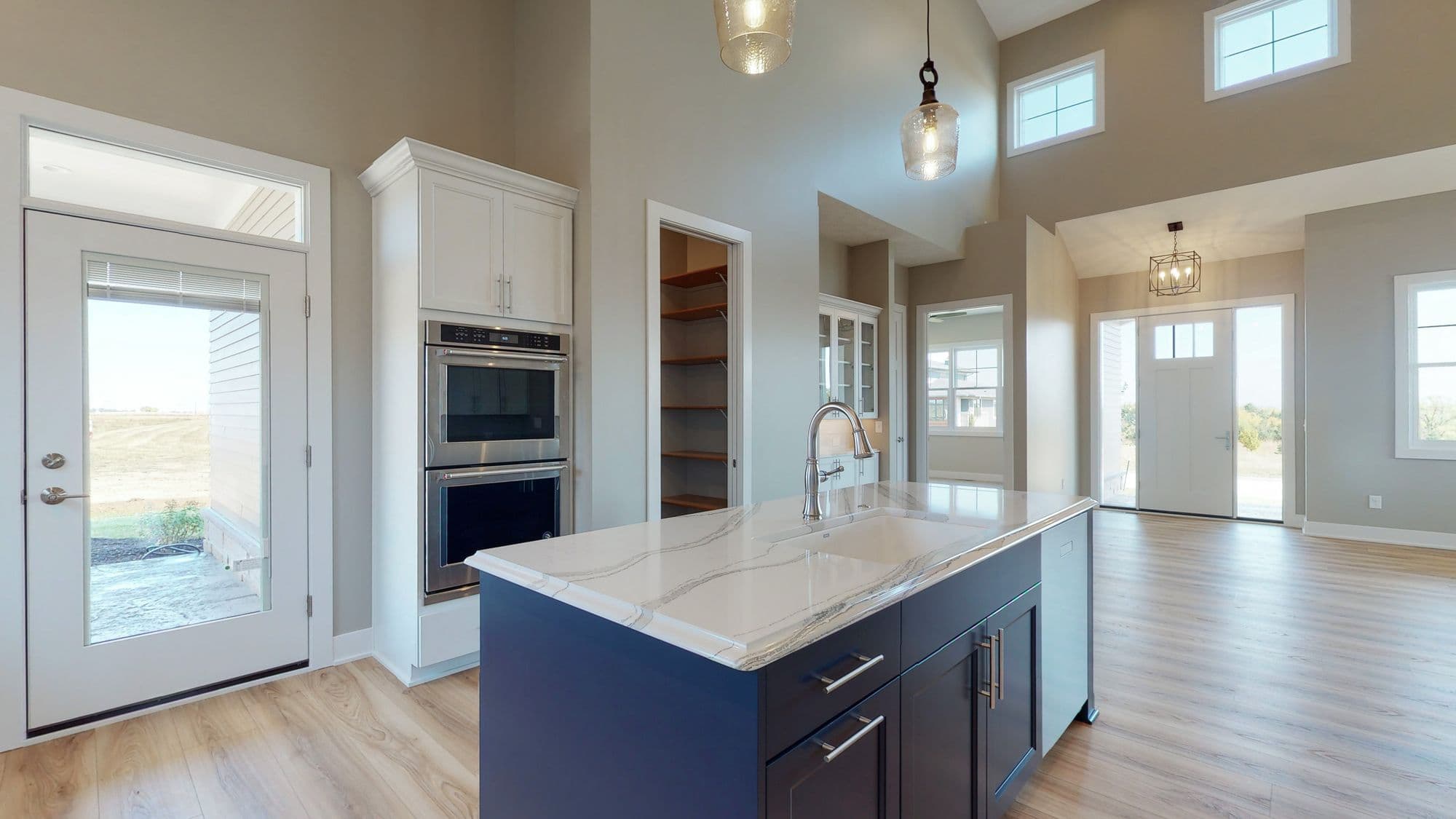 Modern kitchen with blue island and white marble countertops, featuring stainless steel appliances and a pantry. The open floor plan leads to the entryway with a clear view of the front door.