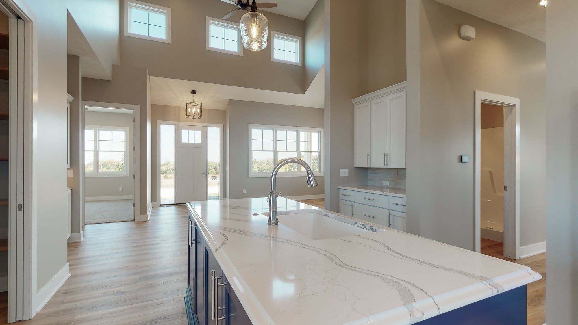 Open-concept kitchen showcases a large marble-topped island with a chrome faucet, accompanied by light-colored cabinetry and a double-height ceiling with transom windows. An open doorway leads to a carpeted room.