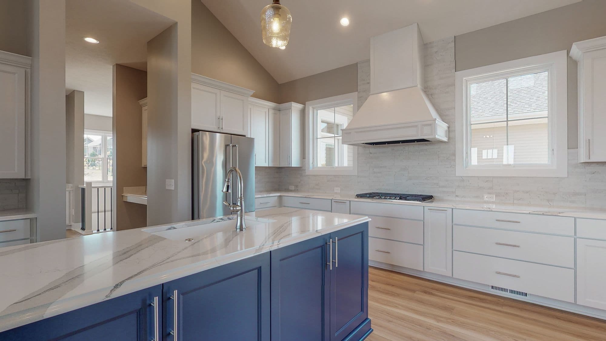 Modern kitchen design featuring white cabinets, marble countertops, and a navy blue island. The space includes stainless steel appliances, a white range hood, and a view into another room.