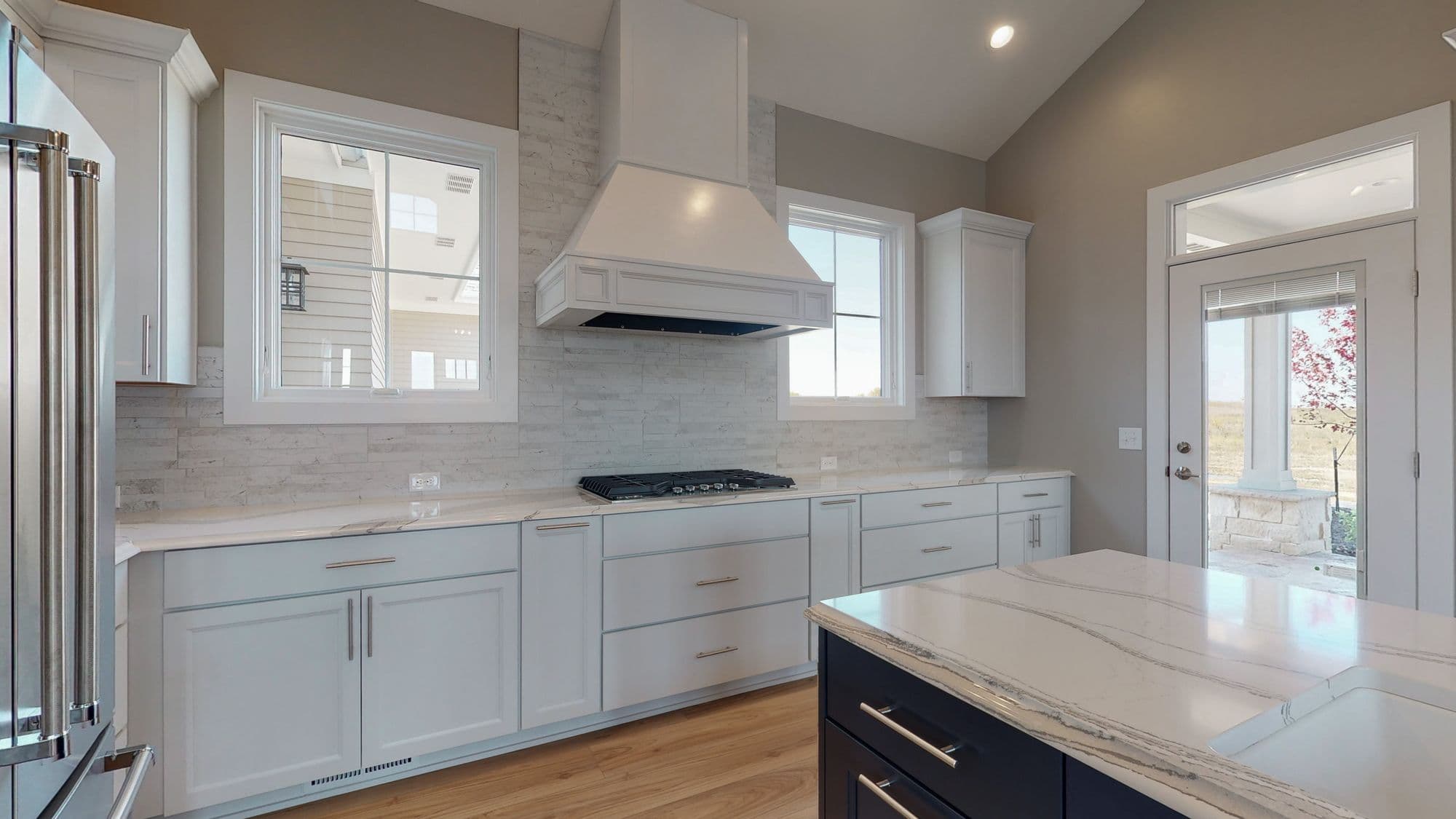 Bright kitchen with white cabinets, stainless steel appliances, and marble countertops and island. A range hood is above the cooktop on the counter, and there are two windows.