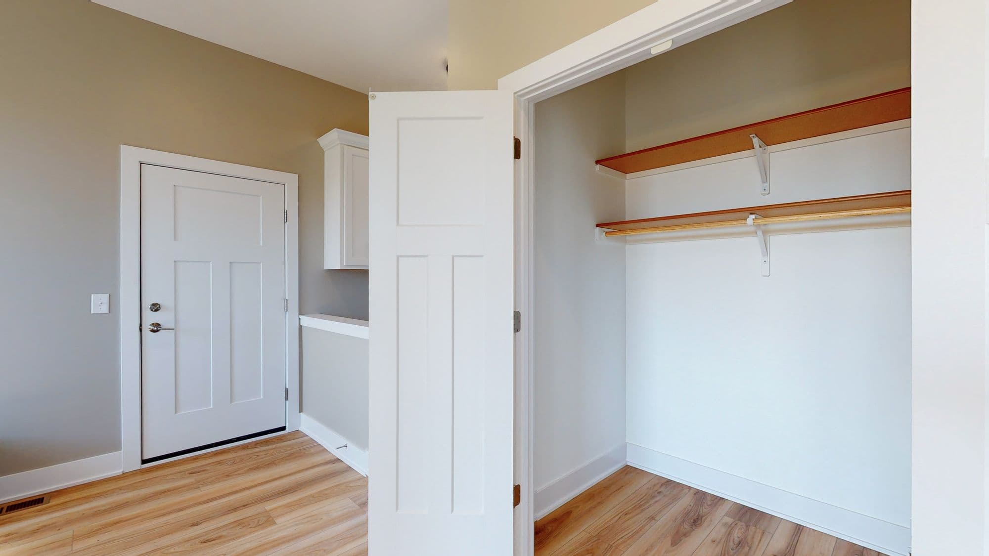 Entryway features a white paneled door leading into the space, alongside an open closet with wood shelving and a rod for hanging clothes. The floors are light wood.