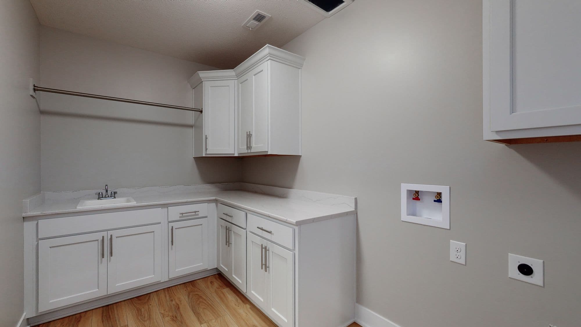 The laundry room features white cabinetry, a white countertop with a sink, and a hanging rod. The walls are painted gray, and the flooring is a warm wood tone.