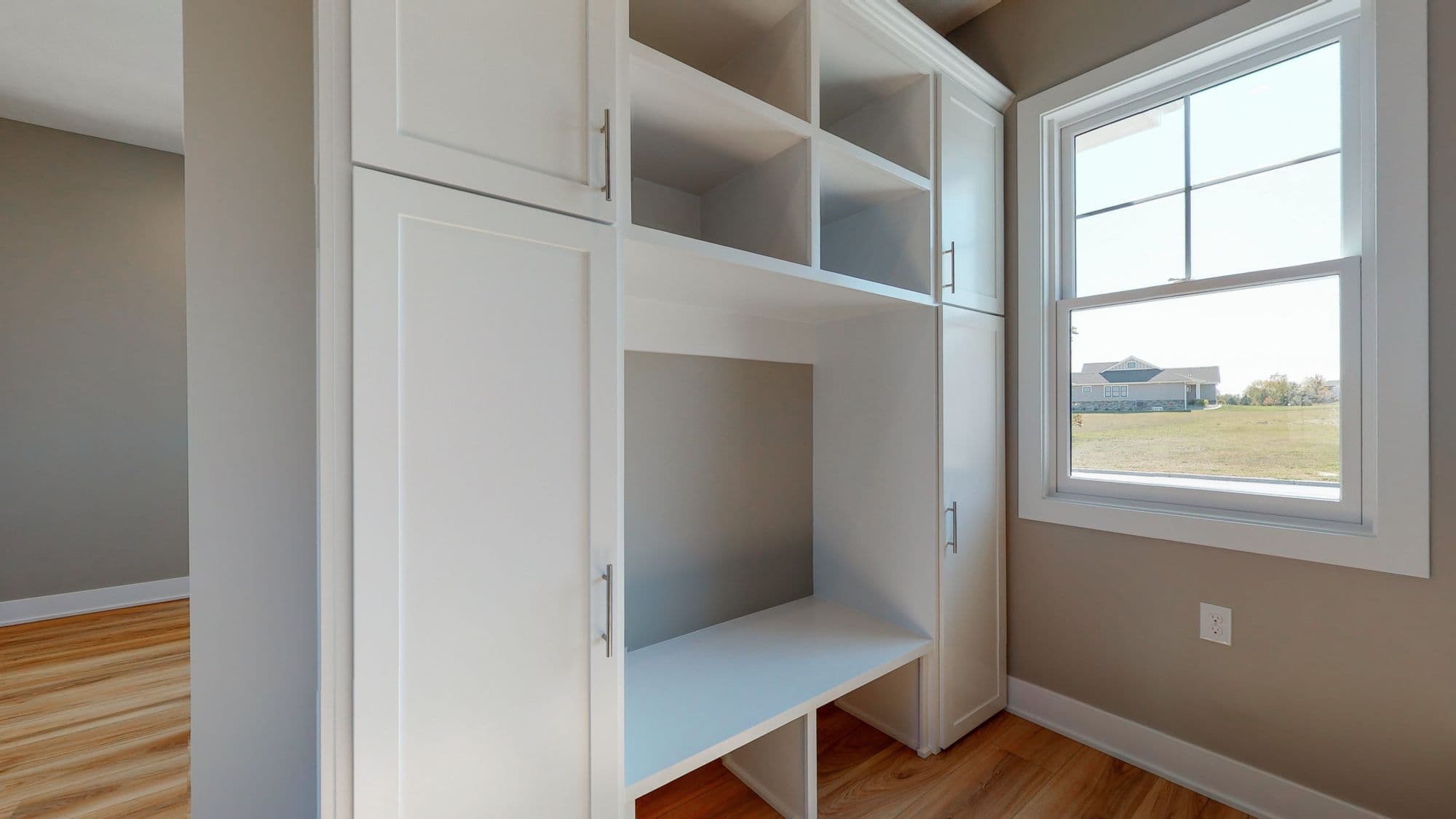 Bright mudroom with built-in white storage cabinets, shelves, and bench. A large window provides natural light and a view of the lawn and nearby house.