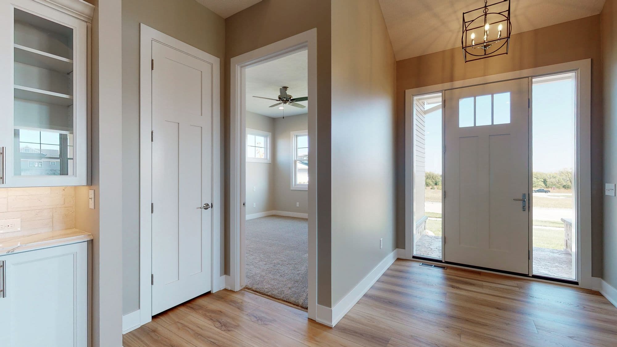 This is a foyer featuring hardwood flooring, a white front door with sidelights, and a view into a bedroom with carpet and windows. The entry is adjacent to a white cabinet and a white door.