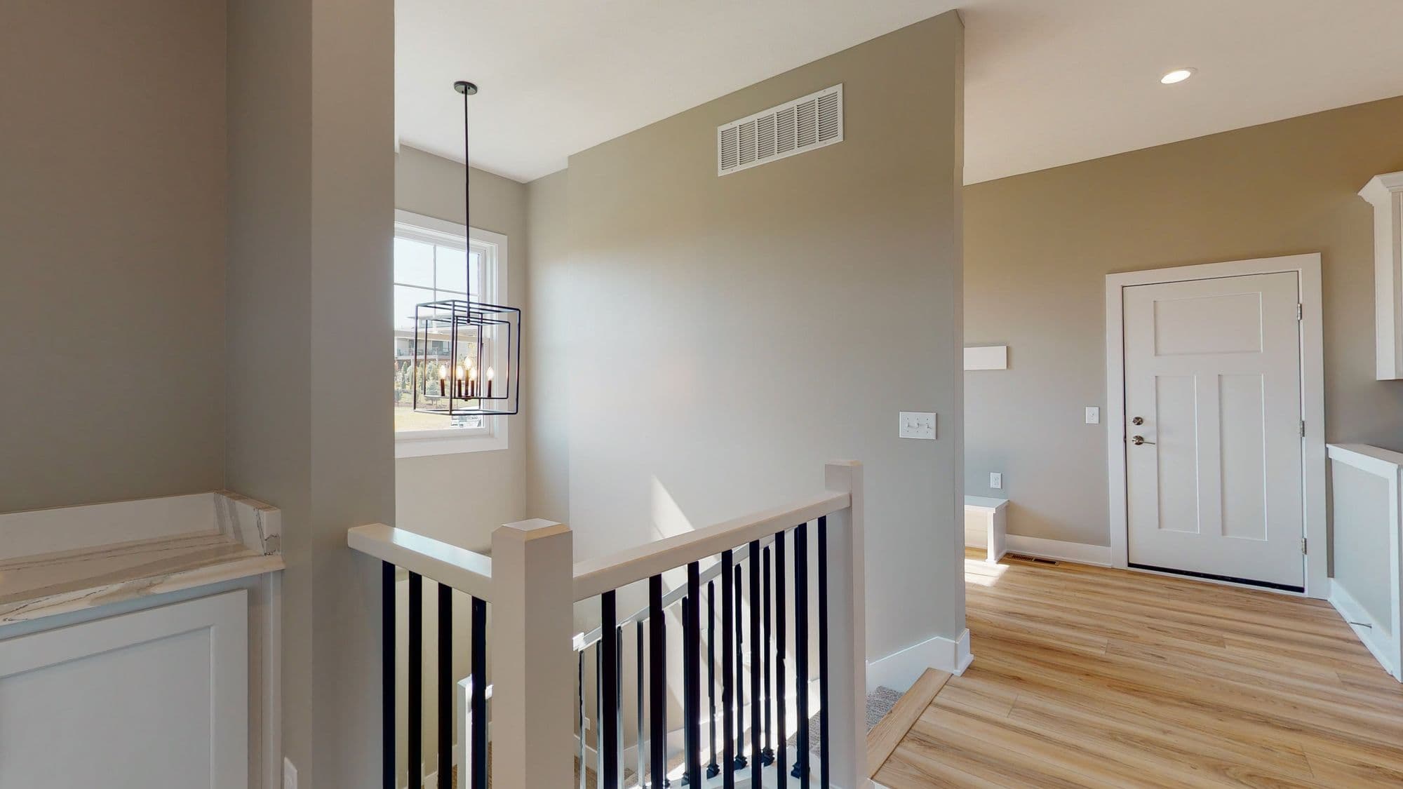 Interior view of a home's upper landing featuring a modern black chandelier, railing with black spindles and white posts, and views into adjacent rooms. The flooring is light wood, and the walls are painted a neutral tone.