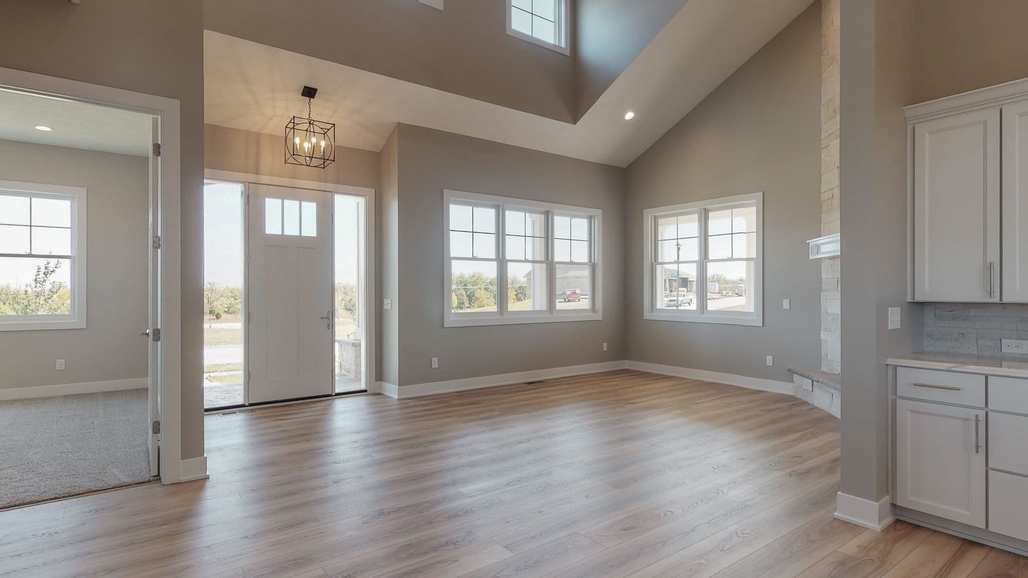 Bright living room with vaulted ceilings and light wood floors. Large windows provide natural light to the space with a modern light fixture hanging near the front door.