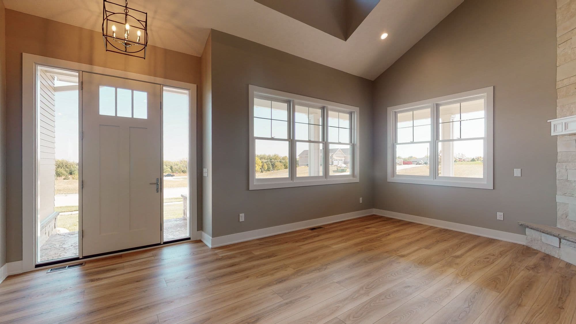 Entryway with open door and sidelights, adjacent to a living room with two windows and hardwood floors. The room features vaulted ceilings and neutral gray walls.