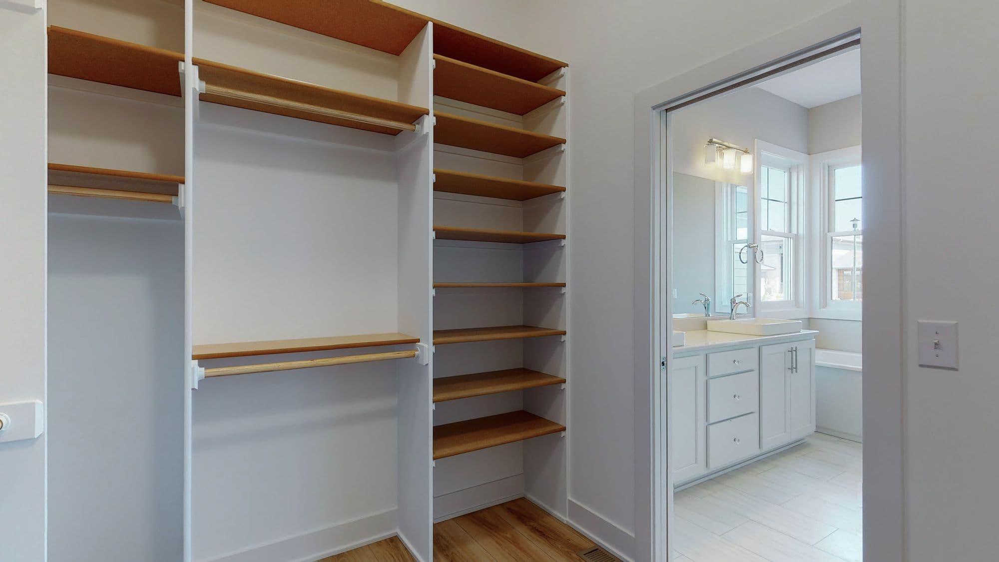 Walk-in closet with built-in shelving and wooden rods, adjacent to a bright bathroom with a vanity and windows. The closet features open storage and shelving, while the bathroom has a modern design.