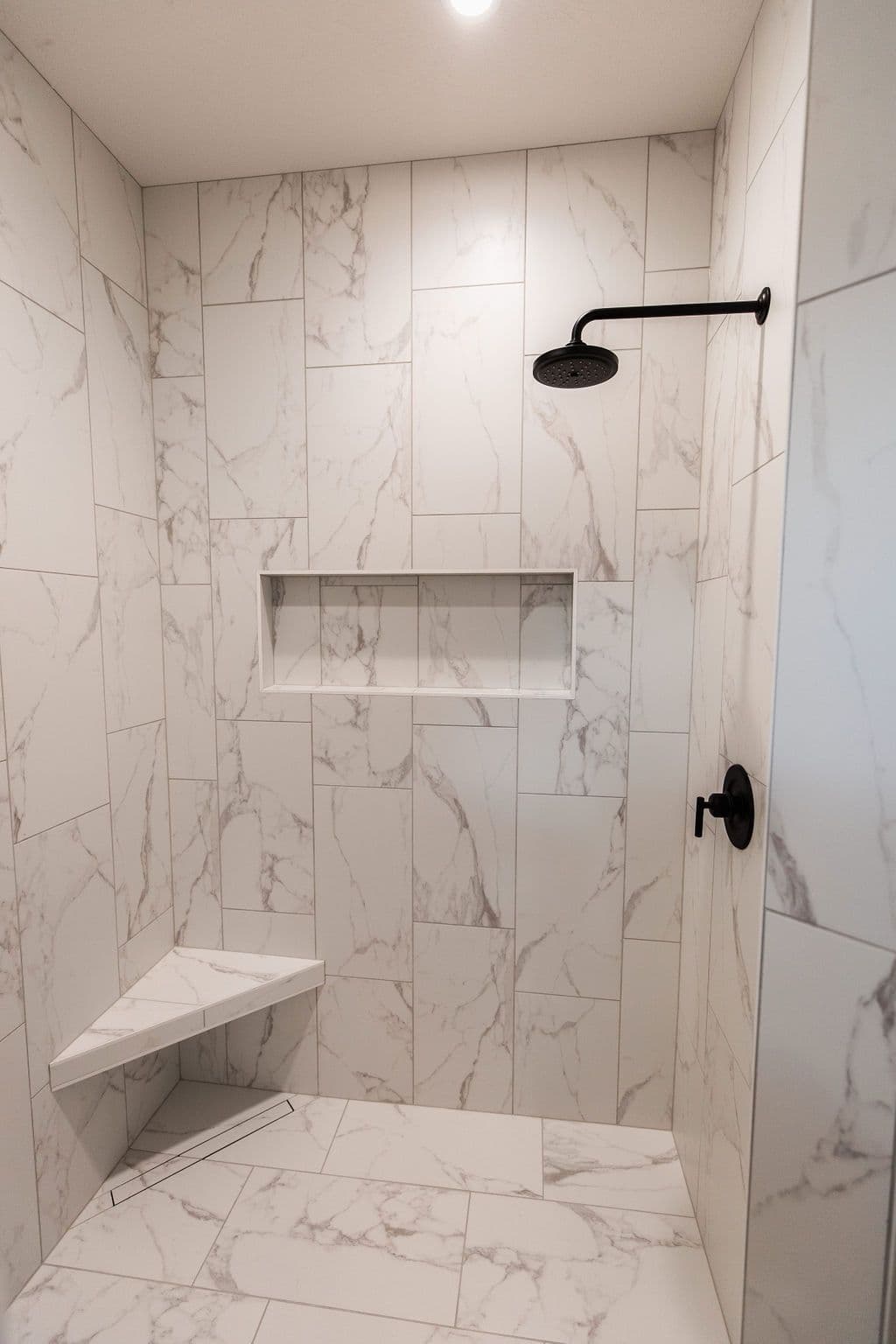 Modern shower with marble-patterned tile walls and floor featuring black showerhead and faucet fixtures. The shower includes a corner seat and recessed niche.