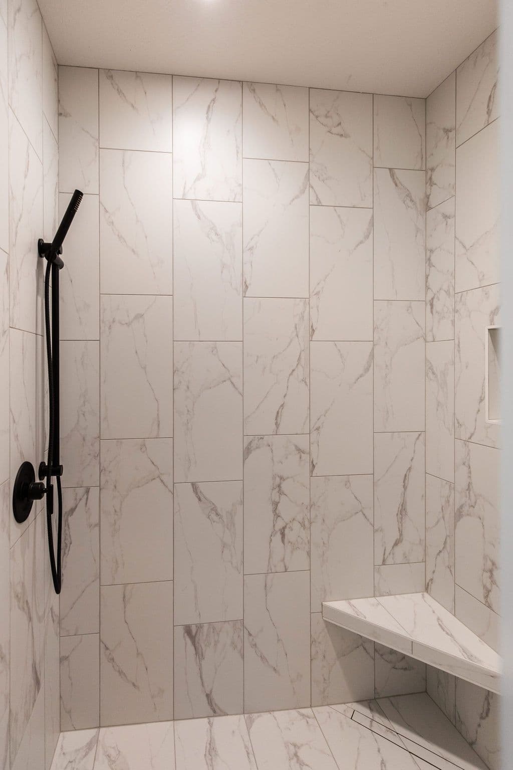 Modern bathroom features a white marble tiled shower with a black shower head fixture and a built-in corner bench. The shower also includes a recessed shower caddy.