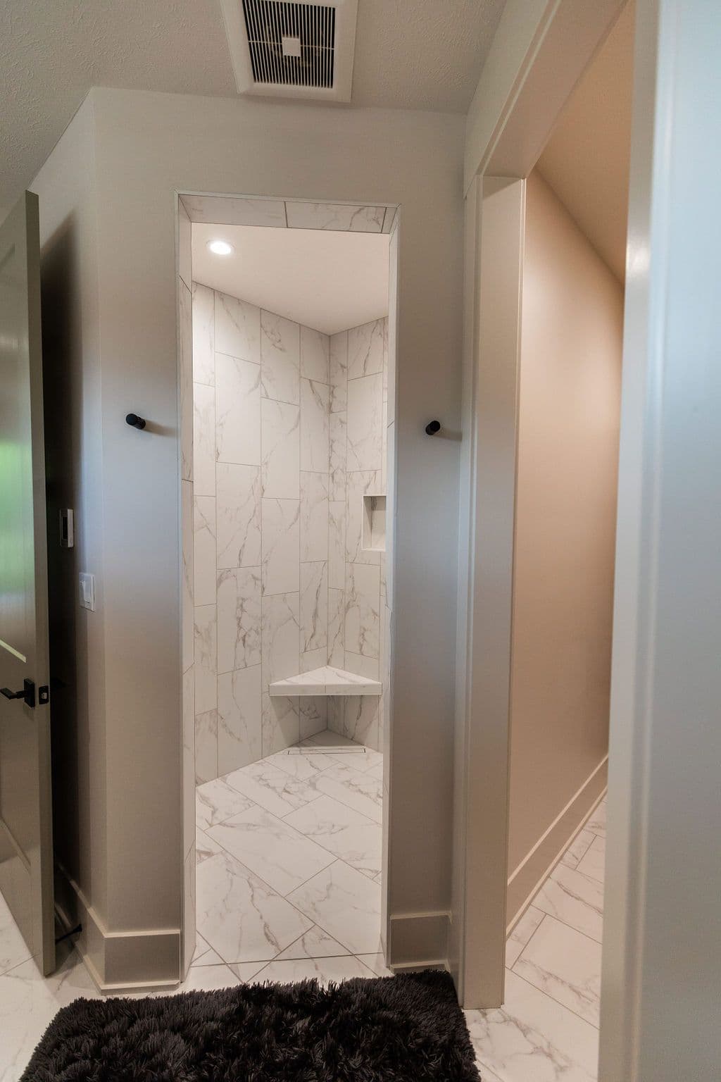 Modern bathroom featuring a walk-in shower with marble tile and a corner bench seat. The bathroom has a dark gray shag rug and a vent fan.