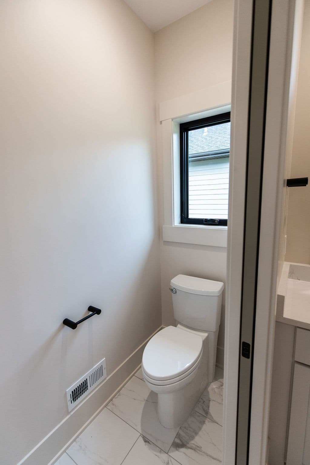 Powder room with white toilet, black toilet paper holder, and a window with black frame. The room has marble tile flooring and light-colored walls.