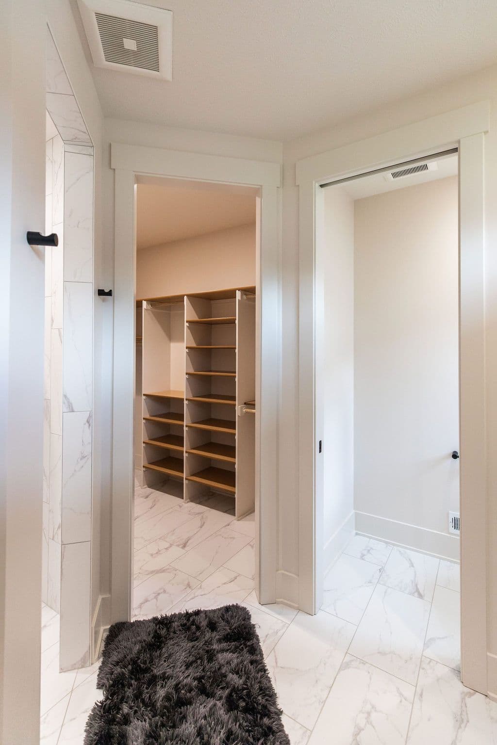 The photo showcases a walk-in closet with built-in shelving. A gray rug is positioned just outside the closet entrance, which has a marble tiled floor.