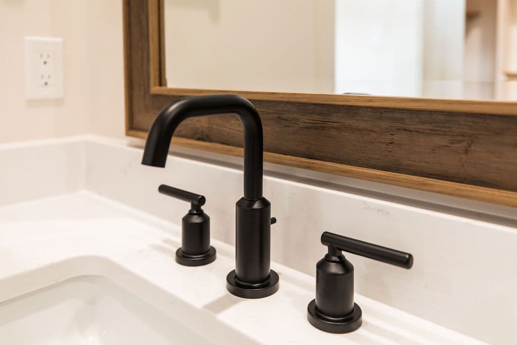 Bathroom features a modern matte black faucet with separate hot and cold water controls, atop a white countertop with integrated sink. A wooden-framed mirror hangs above the sink, and a white electrical outlet is visible on the wall.