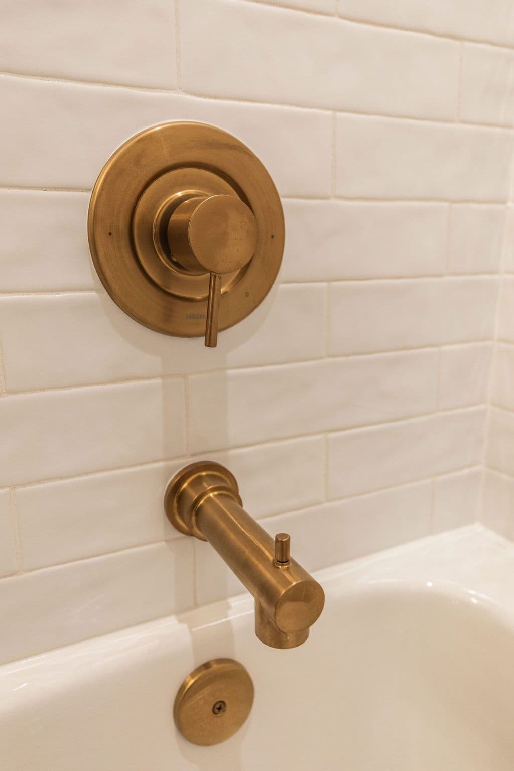 Close-up of a brushed gold shower faucet and handle set mounted on white subway tile in a bathroom setting. The hardware includes a round temperature control and a spout.