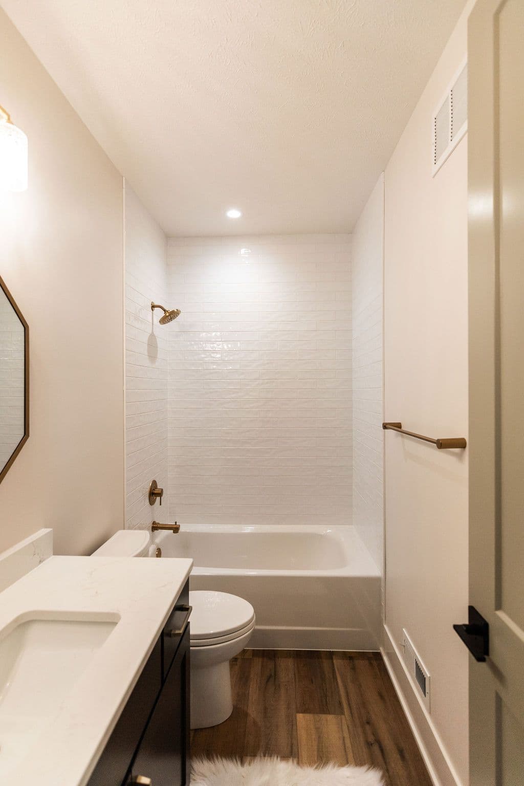 Small bathroom featuring a white subway tile shower, white toilet and sink, dark vanity, and wood-look flooring. Bronze fixtures and a light colored wall paint create a modern and clean aesthetic.