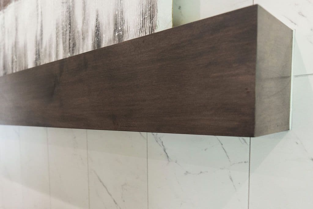 The image shows a floating wood shelf mounted on a white marble tile wall with gray veining. A piece of artwork with gray and white tones hangs above the shelf.