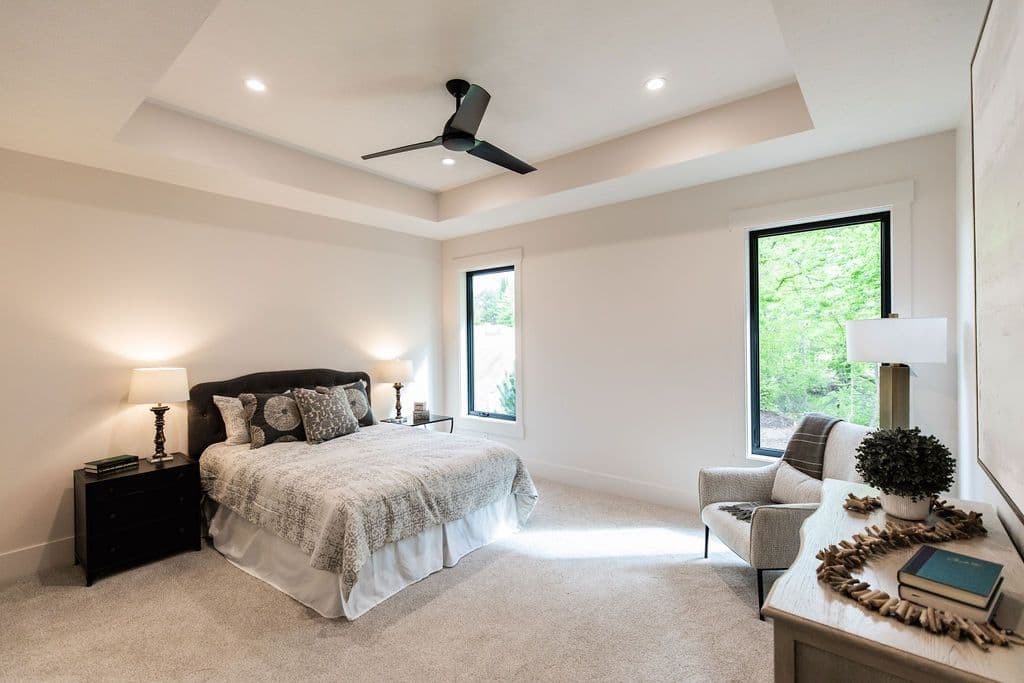 A bright and airy bedroom features neutral carpeting, a dark upholstered bed with patterned bedding, and a black ceiling fan. Two large windows with black frames provide natural light, and a dresser with decorative items sits to the right.