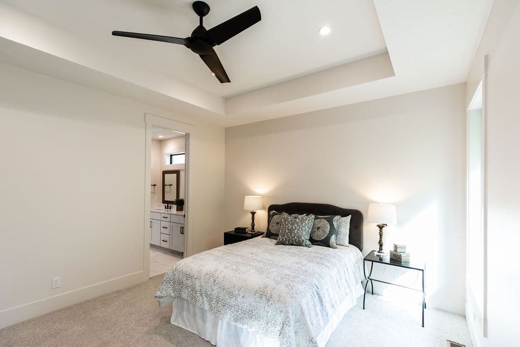 Modern bedroom with neutral decor features a black ceiling fan and an ensuite bathroom visible through a doorway. The room has a bed with decorative pillows, side tables, and is bathed in soft, natural light.