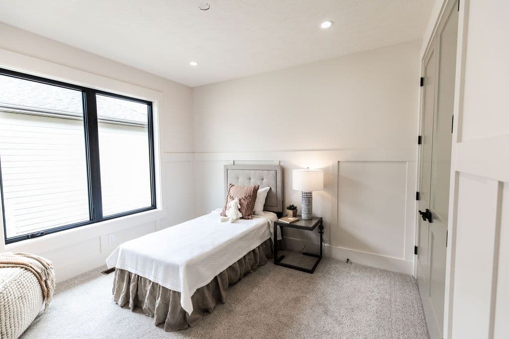 Bright bedroom with a twin bed featuring a tufted gray headboard and a white quilt with a ruffled skirt. A black-framed window and a small nightstand with a lamp are also visible.
