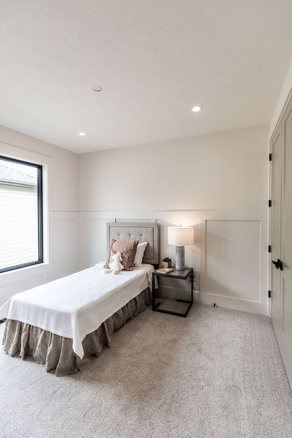 Cozy bedroom features a twin bed with neutral bedding, a tufted headboard, and a small nightstand with a lamp. The room has a soft gray carpet, off-white walls with paneled wainscoting, and a black-framed window.