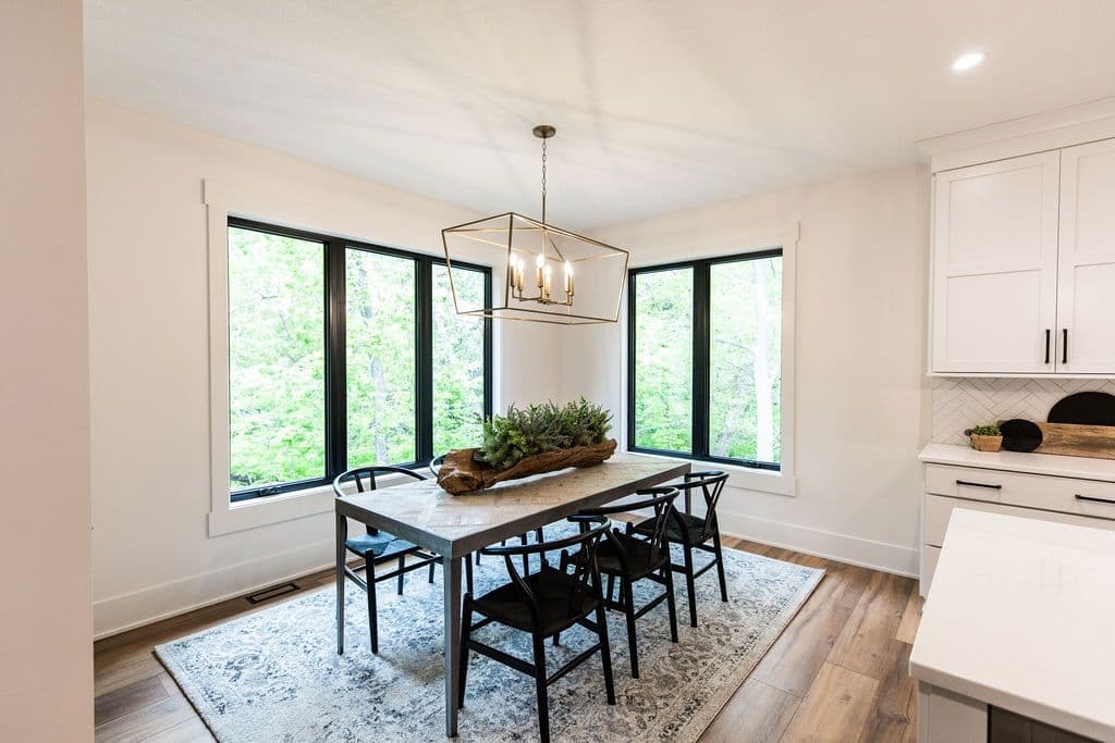 Dining room with a rectangular table and black chairs, large windows with black frames offer ample natural light. A decorative centerpiece sits on the table, and a geometric chandelier hangs above.