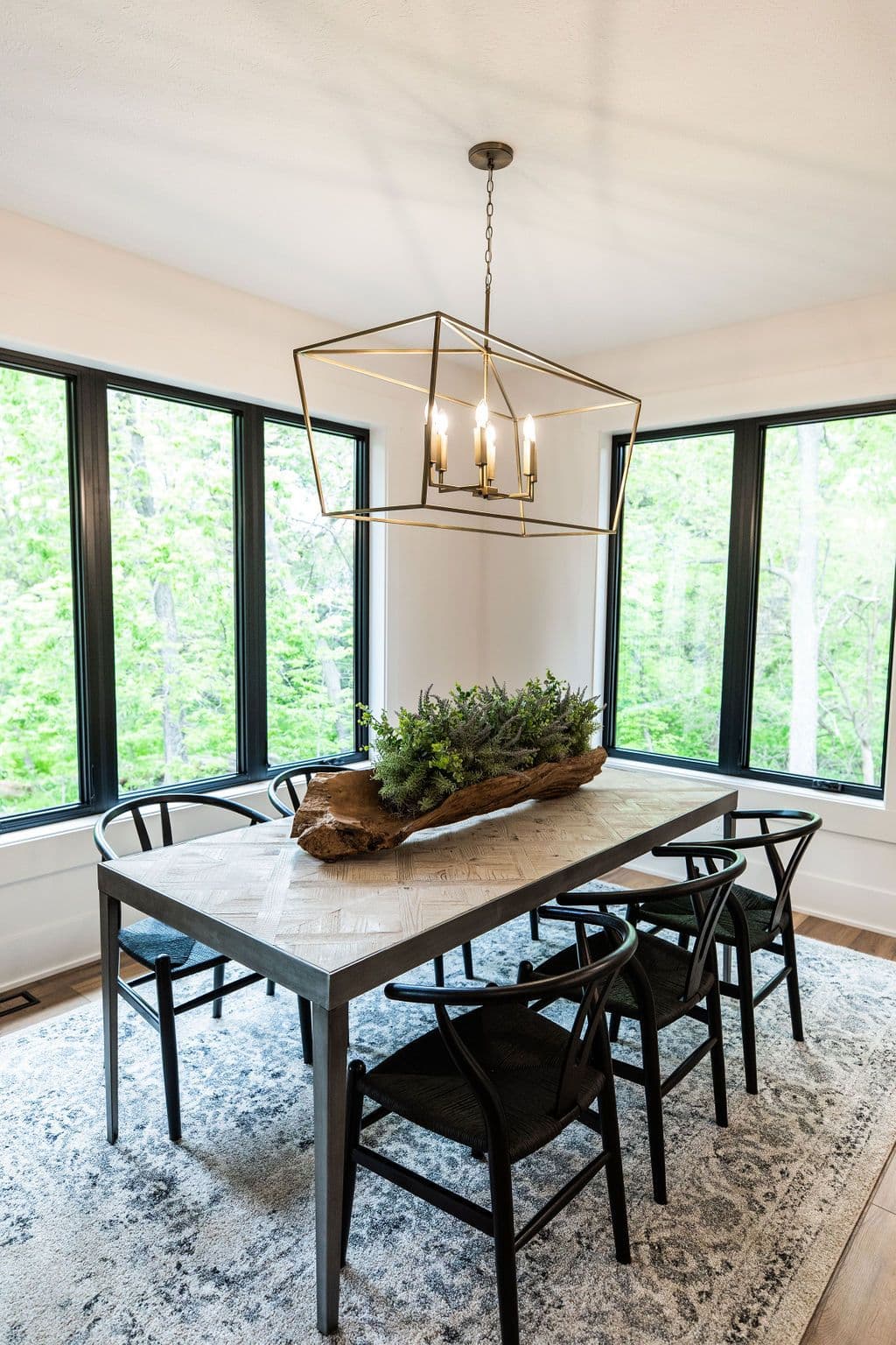 Dining room with a modern chandelier over a herringbone patterned dining table. Black wishbone chairs are arranged around the table, which is topped with a driftwood centerpiece filled with greenery.