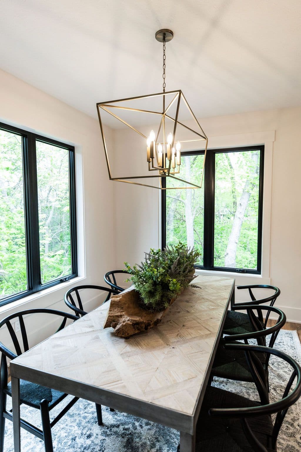 Dining room with light wood table and black wishbone chairs. A modern, geometric chandelier hangs above the table, and large windows with black frames offer views of lush greenery outside.
