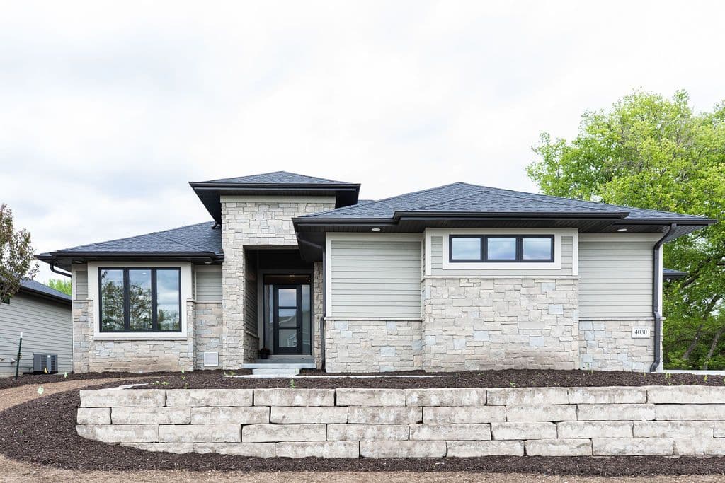 Exterior view of a modern single-story home featuring a combination of stone veneer and horizontal siding. The house has a gabled roof with dark trim, and a retaining wall made of large stone blocks in the front yard.